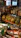 A variety of liquor bottles are displayed on the top shelf, including brands like Johnnie Walker, Gordon’s, and others. Below them, there are several packs of pasta with yellow and green packaging, stacked neatly. The background includes a refrigerated section with drink bottles.