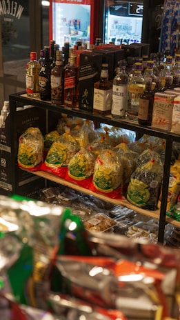 A variety of liquor bottles are displayed on the top shelf, including brands like Johnnie Walker, Gordon’s, and others. Below them, there are several packs of pasta with yellow and green packaging, stacked neatly. The background includes a refrigerated section with drink bottles.