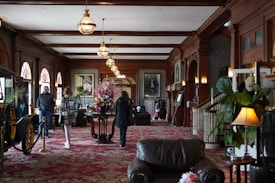 A plush, ornate interior with rich wooden paneling and vintage-style furniture. People are walking through a long corridor adorned with elegant lighting fixtures and elaborate floral arrangements. Large framed portraits line the walls, and the decor suggests an air of historic luxury. A classic car is partially visible to the left, adding to the ambiance.