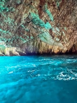 A stunning cave with rugged rock formations inside. The water below is a bright turquoise, reflecting light and creating a vibrant contrast with the earthy browns and greens of the cave walls.