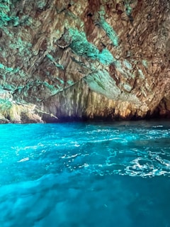 A stunning cave with rugged rock formations inside. The water below is a bright turquoise, reflecting light and creating a vibrant contrast with the earthy browns and greens of the cave walls.