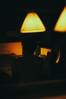 A dimly lit room with warm amber lighting creates a cozy atmosphere. A silhouette of a person is seated at a desk, surrounded by stacks of books and a glass. The overhead lamps emit a soft glow, casting shadows and adding to the intimate setting.