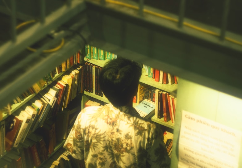 A person in a floral shirt stands in a narrow aisle of a cozy library or bookstore, browsing through a selection of books neatly organized on shelves. The lighting is soft and warm, creating a comfortable and intimate atmosphere. There is a visible sign with text, likely instructions or information for customers.