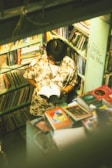 A person with a floral shirt is standing in a small, crowded bookstore or library, reading a book. The shelves are filled with various colorful books, and there is writing on one of the walls. The lighting gives the scene a warm, nostalgic feel.