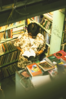 A person with a floral shirt is standing in a small, crowded bookstore or library, reading a book. The shelves are filled with various colorful books, and there is writing on one of the walls. The lighting gives the scene a warm, nostalgic feel.