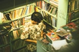 A person stands in a small, cozy bookshop surrounded by shelves filled with an array of books. The individual is wearing a floral shirt and appears to be absorbed in examining something. The room is warmly lit, creating a serene and intimate atmosphere.