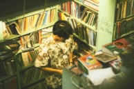A person stands in a small, cozy bookshop surrounded by shelves filled with an array of books. The individual is wearing a floral shirt and appears to be absorbed in examining something. The room is warmly lit, creating a serene and intimate atmosphere.