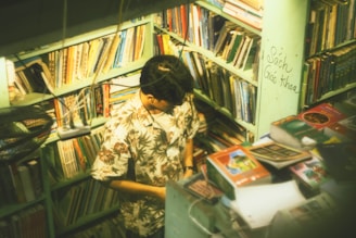 A person stands in a small, cozy bookshop surrounded by shelves filled with an array of books. The individual is wearing a floral shirt and appears to be absorbed in examining something. The room is warmly lit, creating a serene and intimate atmosphere.