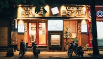 A warmly lit storefront with bold signage featuring Korean characters and the word 'TOJONG'. The establishment appears to be a restaurant, with glowing light boxes displaying menu items. Three parked motorbikes are positioned in front on the dimly lit street. A few decorative plants are visible near the entrance, enhancing the inviting ambiance.