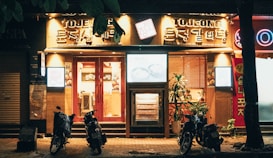 A warmly lit storefront with bold signage featuring Korean characters and the word 'TOJONG'. The establishment appears to be a restaurant, with glowing light boxes displaying menu items. Three parked motorbikes are positioned in front on the dimly lit street. A few decorative plants are visible near the entrance, enhancing the inviting ambiance.