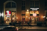 An urban street view with illuminated signs of KFC and Highlands Coffee on a building. People are sitting inside and walking outside, and a white car is parked in front. The atmosphere is lively, with warm lighting creating a cozy ambiance.