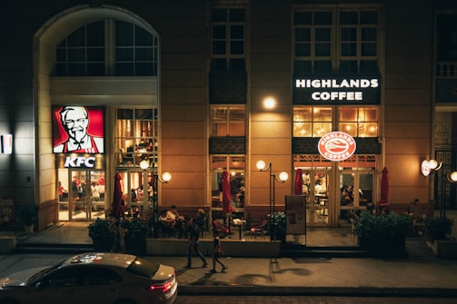An urban street view with illuminated signs of KFC and Highlands Coffee on a building. People are sitting inside and walking outside, and a white car is parked in front. The atmosphere is lively, with warm lighting creating a cozy ambiance.