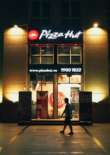 A Pizza Hut restaurant storefront is illuminated at night, with glowing signage and posters on either side of the glass doors. A person walks by in silhouette against the warm lighting of the store's interior, creating a contrast with the dark exterior surroundings.
