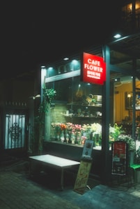 A cozy cafe storefront is illuminated by a soft light, with a selection of flowers displayed in the window. A red sign with 'Cafe Flower' hangs above, and there is a small outdoor bench beside the entrance. The overall ambiance is calm and inviting.
