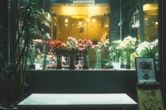 A charming florist display with various colorful flowers in glass vases arranged neatly in a shop window. Warm lighting from inside the store creates a cozy and inviting atmosphere, while plants and decorative elements adorn the exterior. A sign is visible with a vintage aesthetic.
