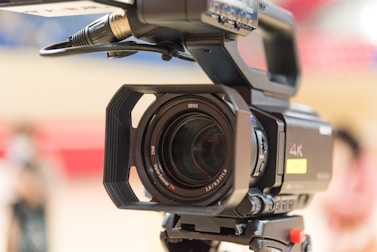 A professional 4K video camera is prominently featured, showcasing the lens with visible branding details. The camera appears mounted on a tripod, hinting at a setup for filming or photography. The background is blurred, emphasizing the camera as the focal point.