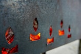 A metallic surface with multiple flame-shaped cutouts, through which a fiery orange-red glow is visible. The metal appears to be rough and weathered, giving the image an industrial feel.