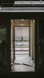 View through the open door of a train, revealing a platform with a sign that says 'Surabaya Gubeng'. The scene includes railway tracks and a person standing in the distance.