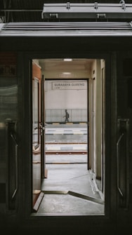 View through the open door of a train, revealing a platform with a sign that says 'Surabaya Gubeng'. The scene includes railway tracks and a person standing in the distance.