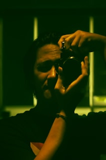 A person is holding a camera up to their eye, appearing to take a photograph. The lighting is low, casting strong shadows across their face. The image has a greenish hue, possibly due to artificial lighting or digital color grading.