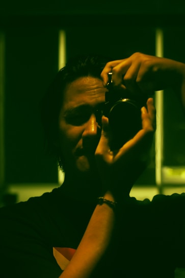 A person is holding a camera up to their eye, appearing to take a photograph. The lighting is low, casting strong shadows across their face. The image has a greenish hue, possibly due to artificial lighting or digital color grading.