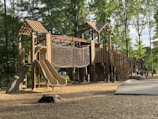 A wooden playground structure with slides, rope climbing net, and multiple levels is set in a park surrounded by tall, leafy trees. The ground is covered with wood chips, and sunlight filters through the trees, casting shadows on the structure. The environment is serene and inviting.