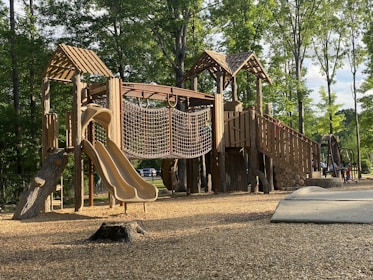 Wooden playground structure with slides and climbing features in a sunny park.