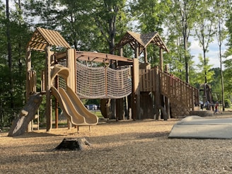 A wooden playground structure with slides, rope climbing net, and multiple levels is set in a park surrounded by tall, leafy trees. The ground is covered with wood chips, and sunlight filters through the trees, casting shadows on the structure. The environment is serene and inviting.