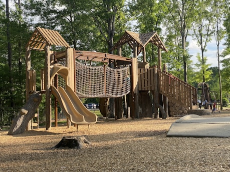 A wooden playground structure with slides, rope climbing net, and multiple levels is set in a park surrounded by tall, leafy trees. The ground is covered with wood chips, and sunlight filters through the trees, casting shadows on the structure. The environment is serene and inviting.
