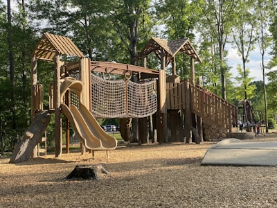 A wooden playground structure with slides, rope climbing net, and multiple levels is set in a park surrounded by tall, leafy trees. The ground is covered with wood chips, and sunlight filters through the trees, casting shadows on the structure. The environment is serene and inviting.