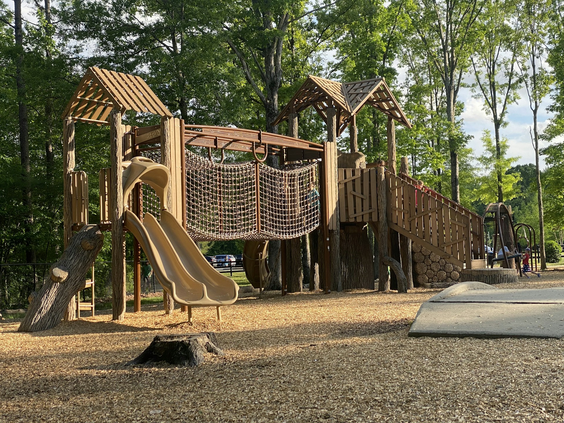A wooden playground structure with slides, rope climbing net, and multiple levels is set in a park surrounded by tall, leafy trees. The ground is covered with wood chips, and sunlight filters through the trees, casting shadows on the structure. The environment is serene and inviting.