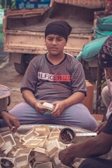 A boy wearing a black turban and a grey T-shirt sits behind a large metal container filled with disposable food trays. His expression is serious as he holds one of the trays in his hands. Surrounding him, another person is seen filling the trays with food using a ladle.