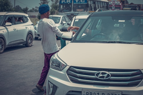 A person wearing a blue turban and white shirt hands an item to someone inside a white Hyundai car. The scene is on a busy road with several cars and buses in the background indicating traffic.