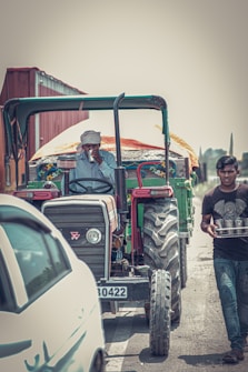 A person wearing a turban is sitting on a tractor, which is part of a rural setting. Another person is walking beside the tractor carrying a tray with several metal cups or glasses. The scene includes a car on the side, and the environment appears dusty with a muted color palette.