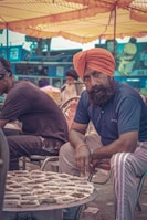 A street vendor dressed in a blue shirt and orange turban sits on a chair, looking directly at the camera, with several trays of dried goods displayed on a table in front of him. The background reveals a busy outdoor market setting with other people and colorful tents providing shade.