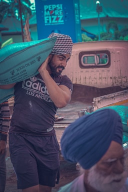 A man wearing a checkered turban is carrying a large green sack on his shoulder. He is dressed in a black Adidas t-shirt and shorts. In the background, there is an old pick-up truck and a billboard with a Pepsi advertisement. Another person wearing a blue turban is visible in the foreground, slightly out of focus.