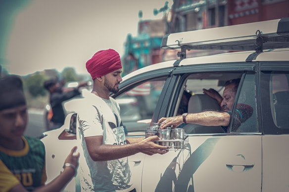 A person wearing a red turban is handing metal containers to another person seated in a white car. The scene seems to be outdoors with a blurred background that includes another person and a vehicle.