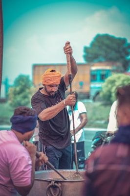 A man wearing an orange turban is stirring a large pot with a long wooden stick. He appears focused on the task as others around him seem engaged in the activity. The background shows blurred trees and a vehicle, contributing to a busy yet communal atmosphere.