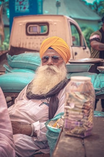 An elderly man with a large white beard and a bright yellow turban is seated outdoors. He is wearing sunglasses and a white shirt with a scarf draped around his shoulders. In front of him is a wooden table with a clear jar of currency notes and a basket of snacks. Behind him, there is a pickup truck loaded with green sacks and the surroundings suggest a market or outdoor setting.
