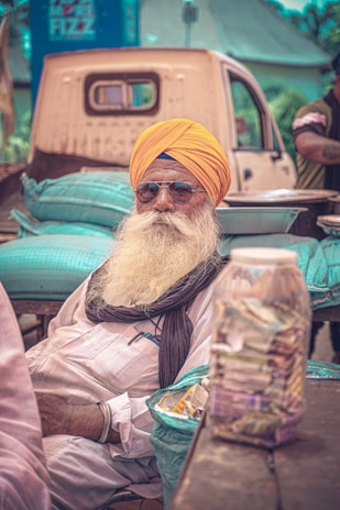 An elderly man with a large white beard and a bright yellow turban is seated outdoors. He is wearing sunglasses and a white shirt with a scarf draped around his shoulders. In front of him is a wooden table with a clear jar of currency notes and a basket of snacks. Behind him, there is a pickup truck loaded with green sacks and the surroundings suggest a market or outdoor setting.