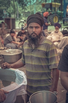 A man wearing a turban and a striped shirt stands by large metal pots, likely involved in outdoor cooking or serving food at a communal event. Several other individuals are seated or working in the background, with trees and a tractor visible.