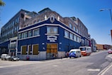 A large corner building painted in dark blue and adorned with stone details is seen rising on a sloping street. The facade features large windows and a prominent sign with 'Edificio Briseas 1949' on it. Several vehicles, including a van and a bus, are parked adjacent to the building, and a few people are gathered at its entrance. The sky is clear and blue, creating a vivid contrast with the building.