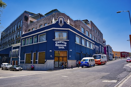 A large corner building painted in dark blue and adorned with stone details is seen rising on a sloping street. The facade features large windows and a prominent sign with 'Edificio Briseas 1949' on it. Several vehicles, including a van and a bus, are parked adjacent to the building, and a few people are gathered at its entrance. The sky is clear and blue, creating a vivid contrast with the building.