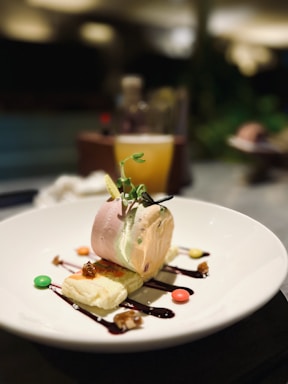 A gourmet dessert consisting of a smooth ice cream scoop placed on a slice of cake, garnished with a small sprig of greenery. The dish is beautifully plated with drizzles of sauce, small nuts, and colorful candies on a white plate that contrasts with the dimly lit background.