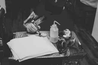 A person with tattoos is sitting at a table, preparing a tattoo gun. The person appears to be focused on their work, with a bottle of ink or cleaning solution and some paper towels on the table. The image is in black and white, giving it a classic and focused atmosphere.