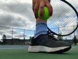 A close-up of a person holding a bright green tennis ball against the strings of a tennis racket. The person is wearing black athletic shoes and striped blue socks while standing on a tennis court. The background includes a cloudy sky and a chain-link fence with trees and buildings in the distance.