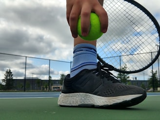 A close-up of a person holding a bright green tennis ball against the strings of a tennis racket. The person is wearing black athletic shoes and striped blue socks while standing on a tennis court. The background includes a cloudy sky and a chain-link fence with trees and buildings in the distance.