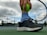 A close-up of a person holding a bright green tennis ball against the strings of a tennis racket. The person is wearing black athletic shoes and striped blue socks while standing on a tennis court. The background includes a cloudy sky and a chain-link fence with trees and buildings in the distance.