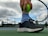 A close-up of a person holding a bright green tennis ball against the strings of a tennis racket. The person is wearing black athletic shoes and striped blue socks while standing on a tennis court. The background includes a cloudy sky and a chain-link fence with trees and buildings in the distance.