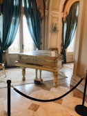 An elegant room with high ceilings and ornate architecture contains a decorative grand piano with intricate designs. The piano is positioned on an ornate rug, surrounded by polished marble flooring. Rich teal curtains hang beside decorative glass doors, and there is a crystal chandelier mounted on the wall.